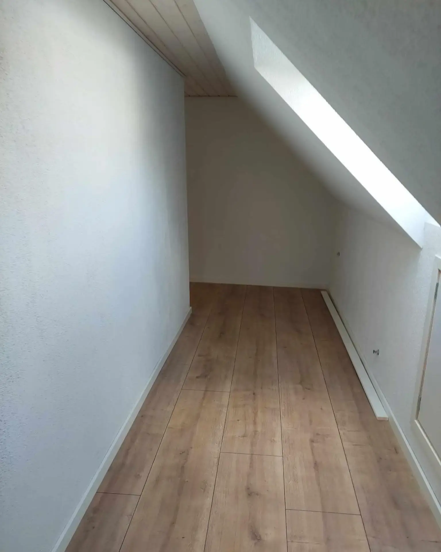 Lille rum under skrå væg med trægulv og hvid væg, perfekt til opbevaring.