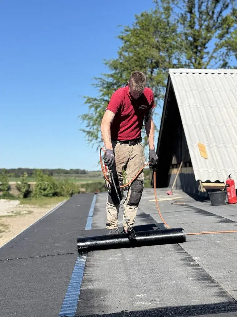 Mand lægger bitumen tagdækning på tag med værktøj i solrigt vejr.