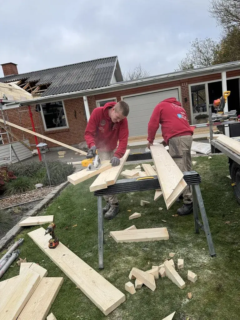 Håndværkere arbejder med træmaterialer på byggeplads foran et hus.