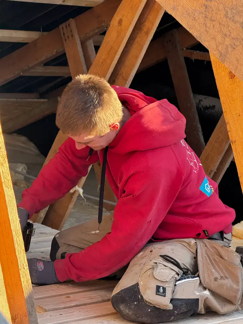 Ung mand i rød hættetrøje arbejder med træ i en byggeplads.